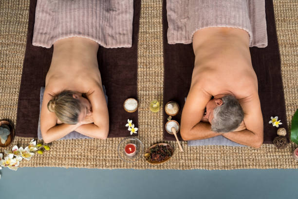 Top view of senior couple lying on front at wellness spa. Mature man and woman lying naked with spa equipment's and flower around them. High angle view of relaxed couple enjoying spa treatment during vacation.